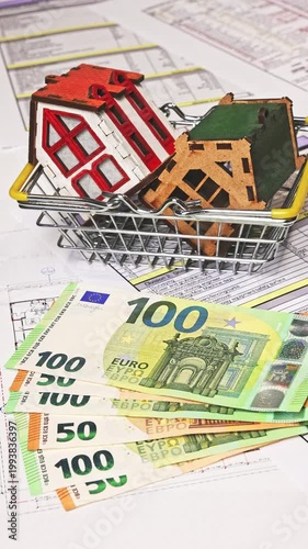 Close-up of house models in a shopping basket while euro banknotes are placed next to keys from a new apartment, symbolizing real estate purchase, ownership, and property investment. 