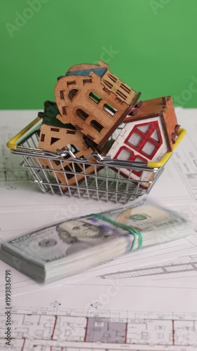 Vertical footage of a stack of US dollar bills being thrown onto architectural blueprints where a shopping basket with miniature house models is already placed, filmed on chroma key. Concept of real