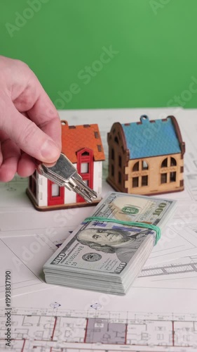 Vertical footage of a hand placing apartment keys on a stack of US dollar bills lying on architectural blueprints with two miniature house models, filmed on chroma key. Concept for real estate