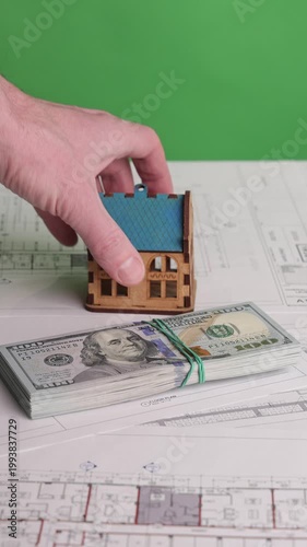 Vertical footage of a hand placing a miniature house with a blue roof behind a stack of US dollar bills on architectural blueprints, filmed on green screen. Concept of real estate investment, property