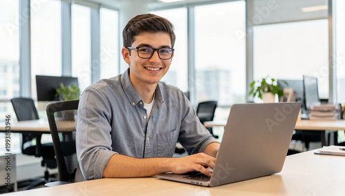 Young man smiles while working on laptop. Glasses and casual shirt complete his look. Office setting with natural light and desks. Professional yet relaxed atmosphere