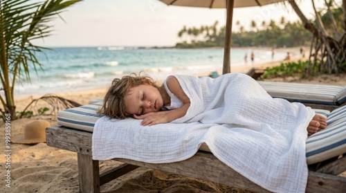 A child rests calmly on a beach lounge, wrapped in a blanket, by the ocean with palm trees and visitors in the background.