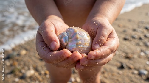 A child's hands cradle a glossy seashell, surrounded by grains of sand, capturing a moment of beach exploration under sunlight.