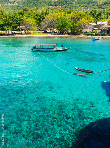 Beautiful tropical atmosphere of Atauro beach in Timor Leste, featuring fishing boats floating on crystal clear turquoise waters where children swim and play.