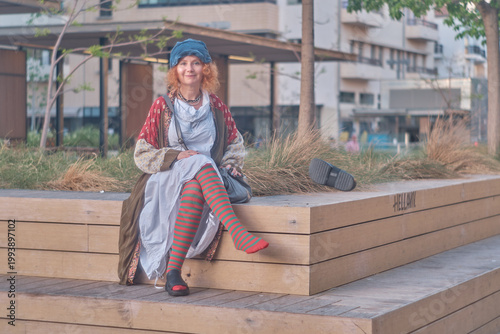 Redheaded woman in colorful bohemian outfit making silence gesture on city bench