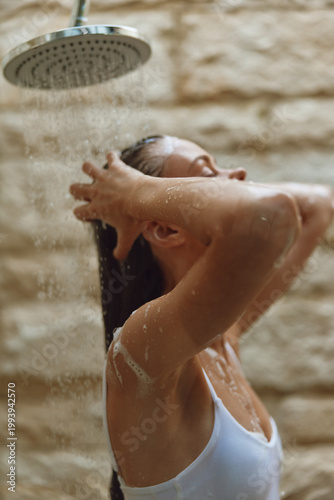 woman showering with eyes closed, enjoying warm water, wet hair, natural light, intimate moment in bathroom with textured wall background