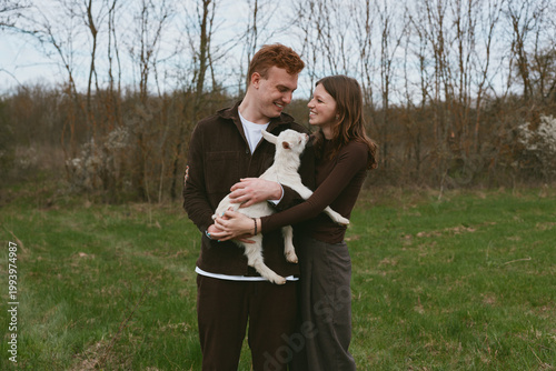 Couple outdoors warmly interacts with a goat kid in a grassy meadow. Happy, affectionate moment captured in natural light, conveying lifestyle, rural living, and animal companionship.