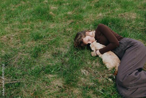 A woman lies on grass embracing a young goat kid in a peaceful outdoor scene. Warm light, relaxed mood, countryside ambiance, gentle companionship, natural textures convey a lifestyle moment of