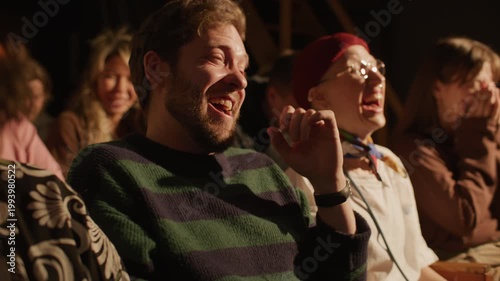 Happy young man and other audience members laughing loudly and reacting to jokes during live stand up show at comedy club