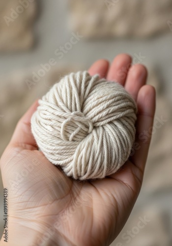 A human hand gently holds a small, perfectly wound ball of natural white cotton yarn against a soft, blurred background.