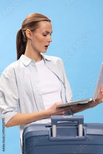 Fotografie Surprised woman with laptop stands behind blue suitcase on blue background