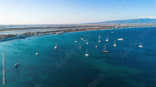 Aerial view of sailboats at anchor in the turquoise water of Poetto beach with salt pans and mountains in the background, Cagliari, Sardinia, Italy