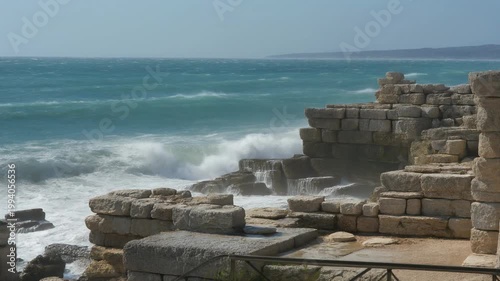 Waves crash against ancient stone ruins on a rugged coastline near a vast sea on a sunny day with clear blue sky