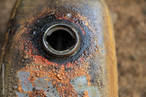 Close-up of a rusty vintage motorcycle fuel tank with peeling paint and weathered texture, showing an open gas filler neck. Grunge industrial background depicting aged metal corrosion and decay.