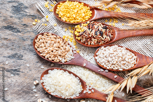 Top view of assorted grains including rice, barley, and mungbean displayed in wooden spoons on a rustic table. Surrounded by dried cereal ears and a linen cloth.