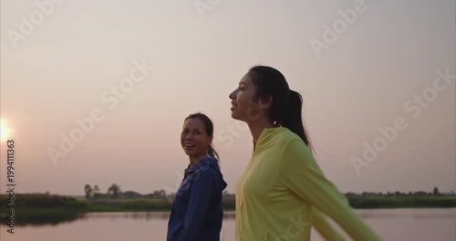 Happy Asian mother and teenage daughter strolling and chatting by lake, authentic family bonding in evening golden light