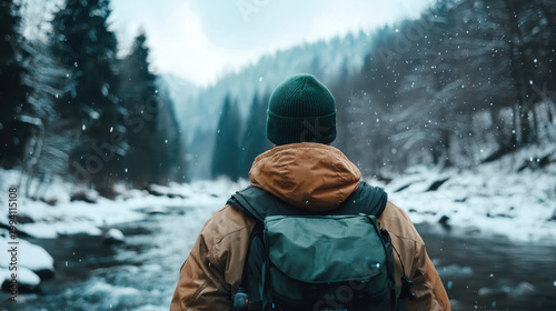  person in a green beanie and brown jacket with a backpack stands by a snowy river surrounded by pine trees and snow-covered mountains.
