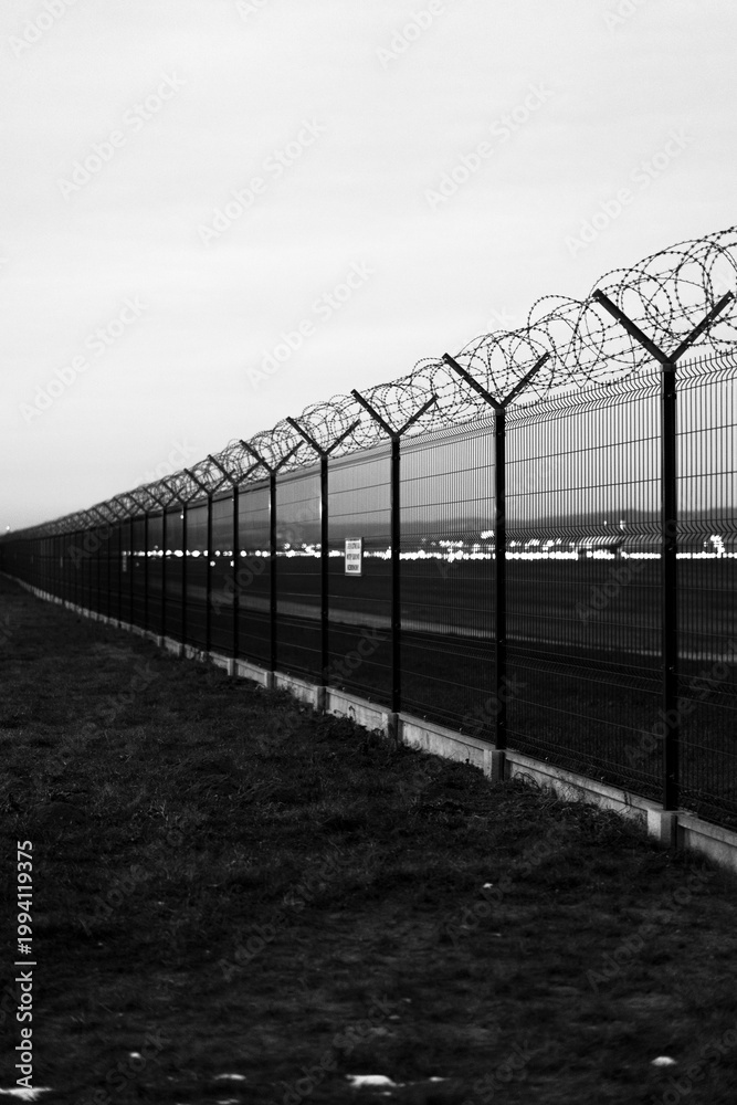 Fototapeta premium Dramatic black and white vertical shot of a high-security metal fence topped with razor wire coils, bordering a restricted airport runway area under a moody, overcast evening sky.