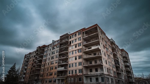 A large abandoned apartment building stands under a stormy sky with dark clouds.