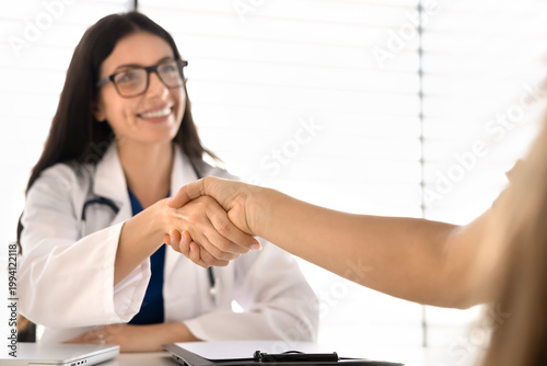 Closeup female doctor sitting at desk shaking hands with woman patient, finalizing successful medical consultation in clinic office. Agreement, professional healthcare services, trust and reliability