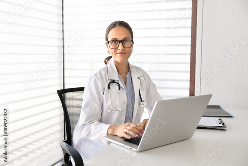 Portrait of smiling female doctor, looking at camera, representing remote medical consultation, professional healthcare services, sitting at desk with laptop in clinic office. Patient care, technology