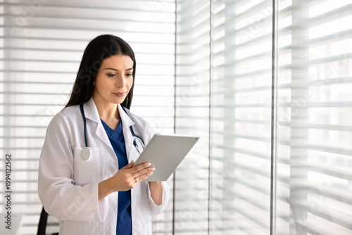 Female doctor reviewing medical online records information, consulting patients remotely using telemedicine application, working on treatment on digital tablet standing in hospital office workplace