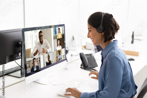 Businesswoman wear headset, participate in online meeting at office desk, communicate with diverse colleagues on computer screen, engage in business work briefing discussion via video call application