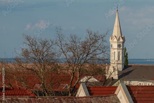 Church tower rising above rooftops in Morbisch, Austria, with leafless trees and Lake Neusiedl in the background, capturing historic village charm, calm atmosphere and scenic countryside view.