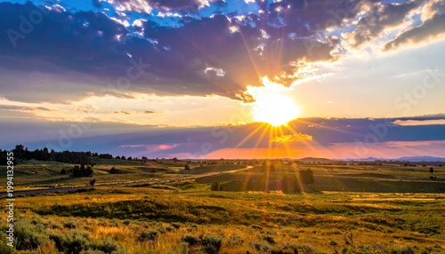 Golden sunset rays burst through dramatic, cloud-filled sky over a grassy plain and road