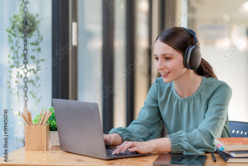 Smiling young businesswoman wearing headphones working on laptop in modern office.