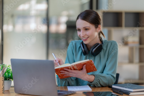 Young businesswoman wearing headset taking notes during online meeting in office.