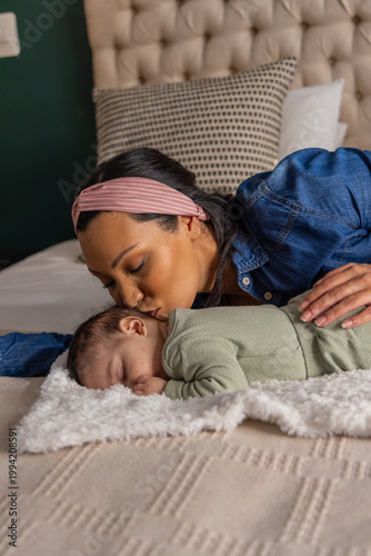 Kissing mother leaning over sleeping infant on bed, wearing denim shirt and pink striped headband