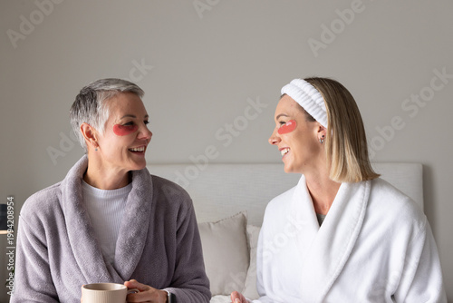 Senior and adult female friends sitting in bedroom wearing plush robes with red under-eye patches