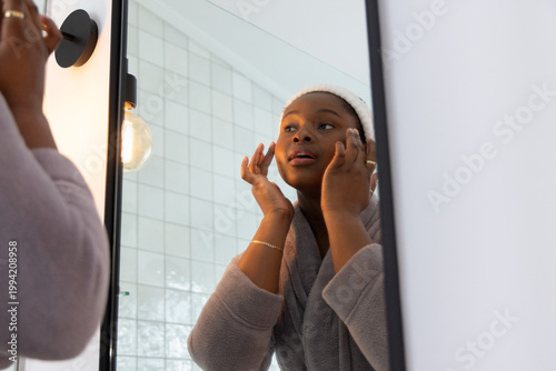 African American woman looking into mirror applying skincare at vanity wearing robe and headband