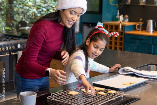 Asian mother in white hat guiding daughter placing star cookies on cooling rack at kitchen island
