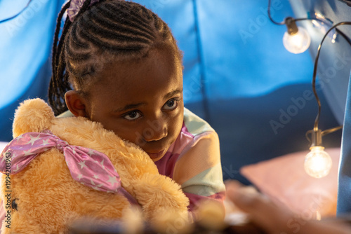 African American girl sitting in blue fort hugging teddy bear, string lights glowing nearby