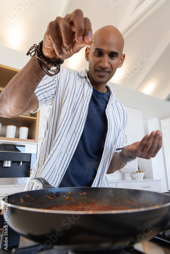 Indian man cooking tomato sauce in kitchen, wearing striped shirt, seasoning skillet with spoon