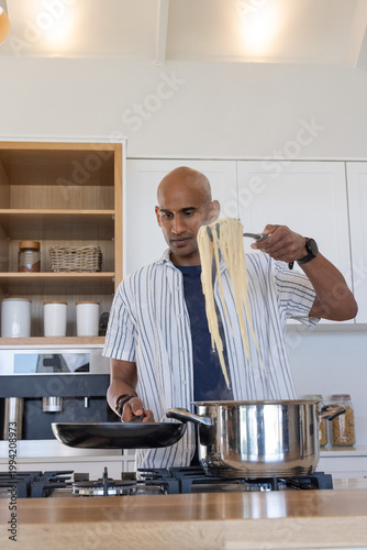 Indian man lifting pasta strands with tongs from stainless pot into frying pan in modern kitchen