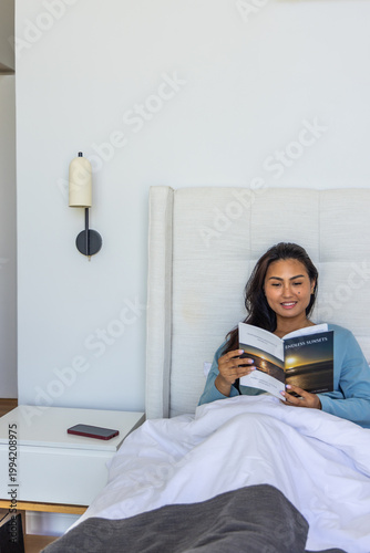 Adult Asian woman sitting in bed reading Endless Sunsets book with phone on nightstand, copy space