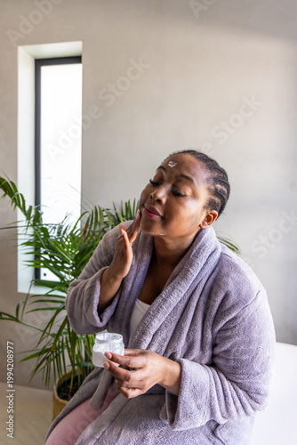 Middle-aged African American woman applying facial cream from open jar in bathrobe in bathroom