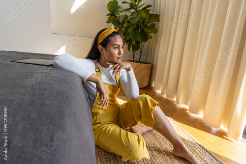 Asian woman sitting on woven rug near sofa in mustard overalls, gazing through curtains with tablet