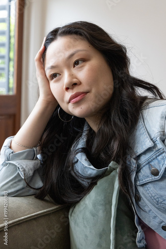 Asian woman leaning on left hand on sofa arm, wearing denim jacket and hoop earrings