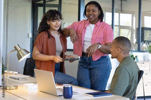 Diverse coworkers standing and gesturing around wooden desk at modern office with laptop tablet