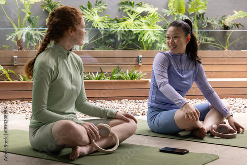 Diverse female friends sitting on yoga mats on patio, in sporty long sleeves, shorts, pilates rings