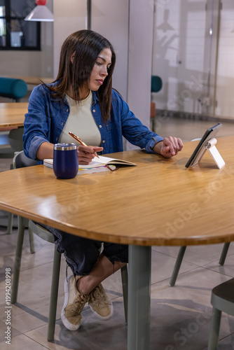 Female sitting at coworking table writing in notebook, glancing at tablet, holding pen, denim shirt