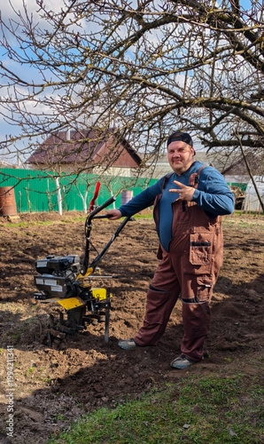 Man in workwear standing by a walk-behind tractor on freshly tilled garden soil, preparing planting beds for spring crops in a sunny, rural home-garden setting.