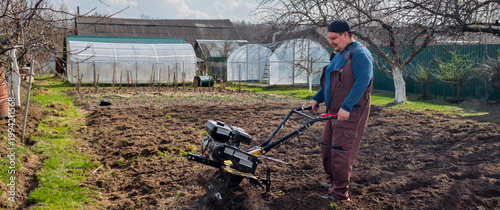 Man working in the garden, operating a walk-behind tractor to cultivate the soil. Greenhouses are visible in the background, showing a scene of active farm work and preparing the ground for planting.