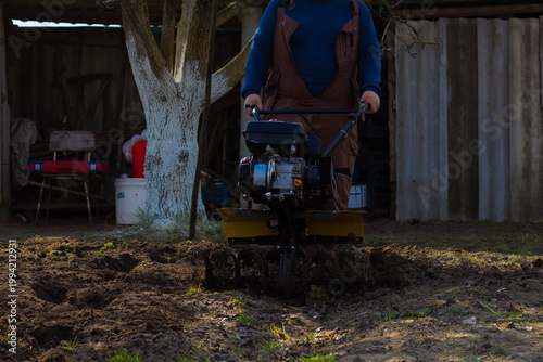 Man operating a walk-behind tractor tilling rich black soil in a home garden, preparing ground for planting and cultivating crops during spring seasonal farm work.