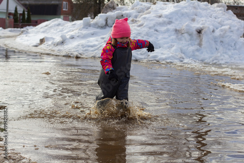 Little girl wearing warm clothes and a pink hat joyfully splashing in a large muddy puddle formed by melting snow, embodying the innocence and fun of childhood during early spring.