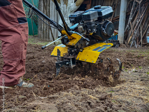 Man operating a walk-behind tractor to till backyard garden soil, preparing and cultivating ground for planting vegetables in a rural home garden setting during spring.